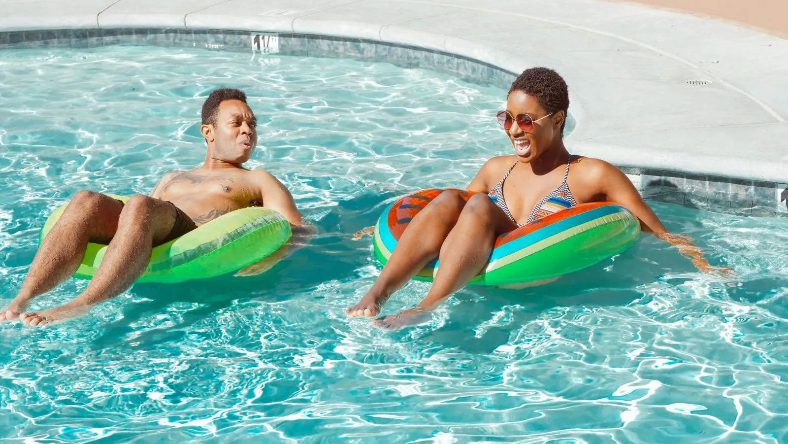 Two friends relaxing on colourful swim rings in a sunny pool