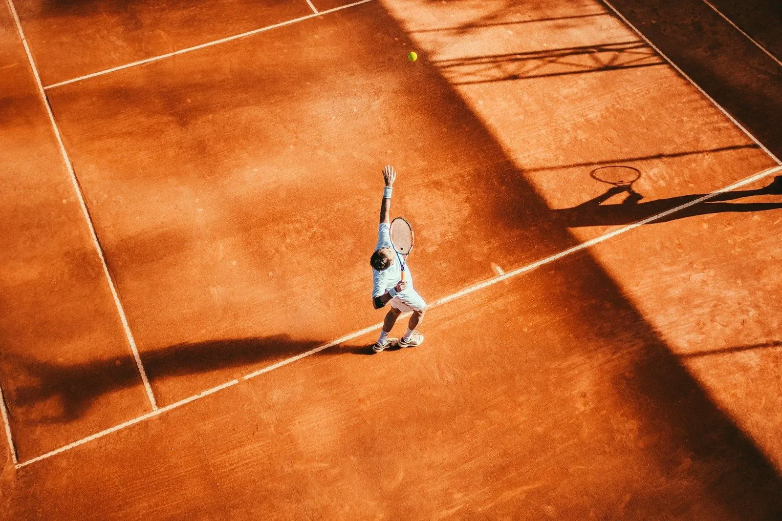 Overhead view of a tennis player serving on a warm orange clay court with long shadows